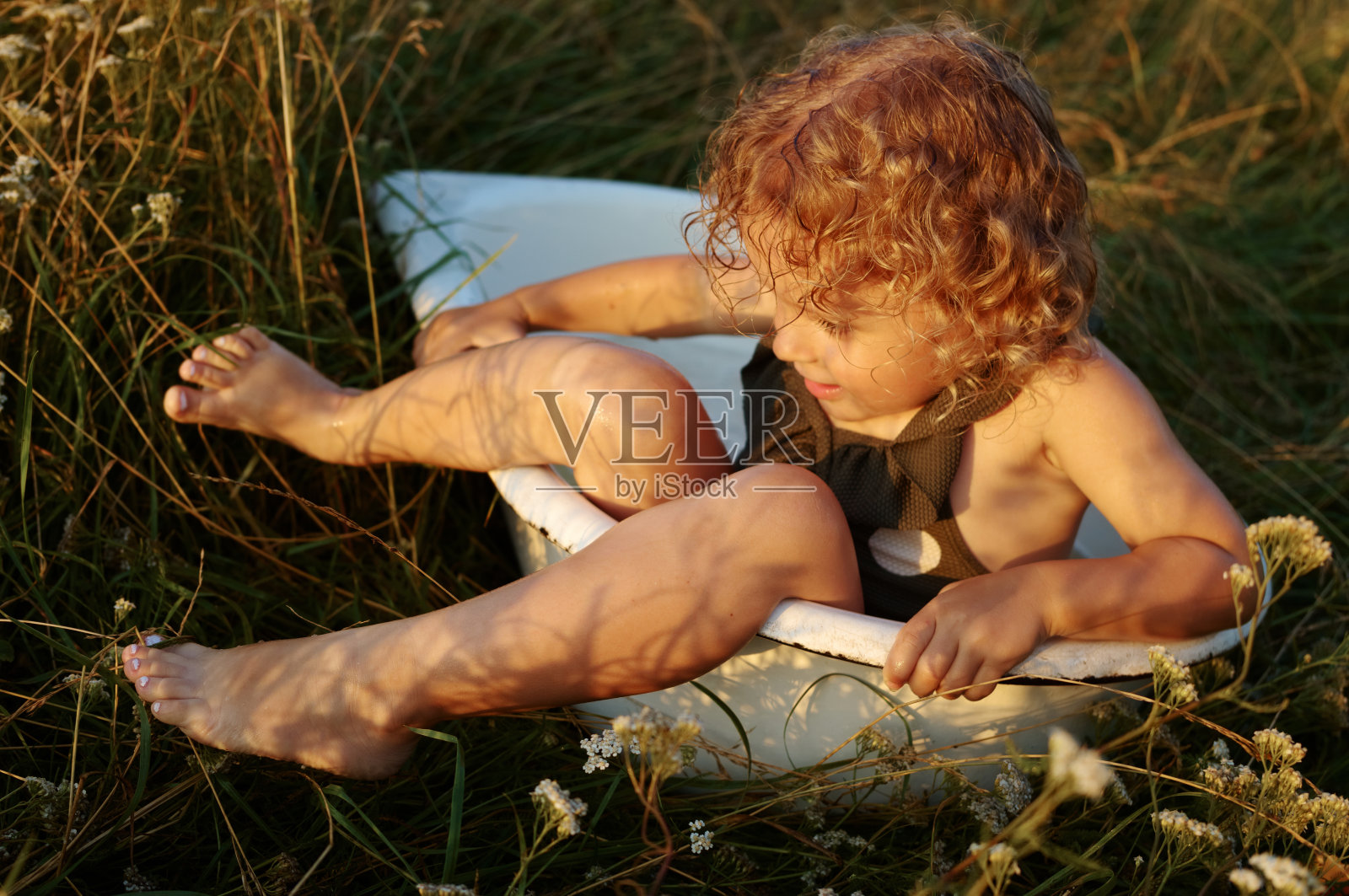 年轻的白种人女孩,波浪状的头发,穿着圆点的泳衣,坐在盛开的草地上洗澡,在温暖的阳光下玩水照片摄影图片