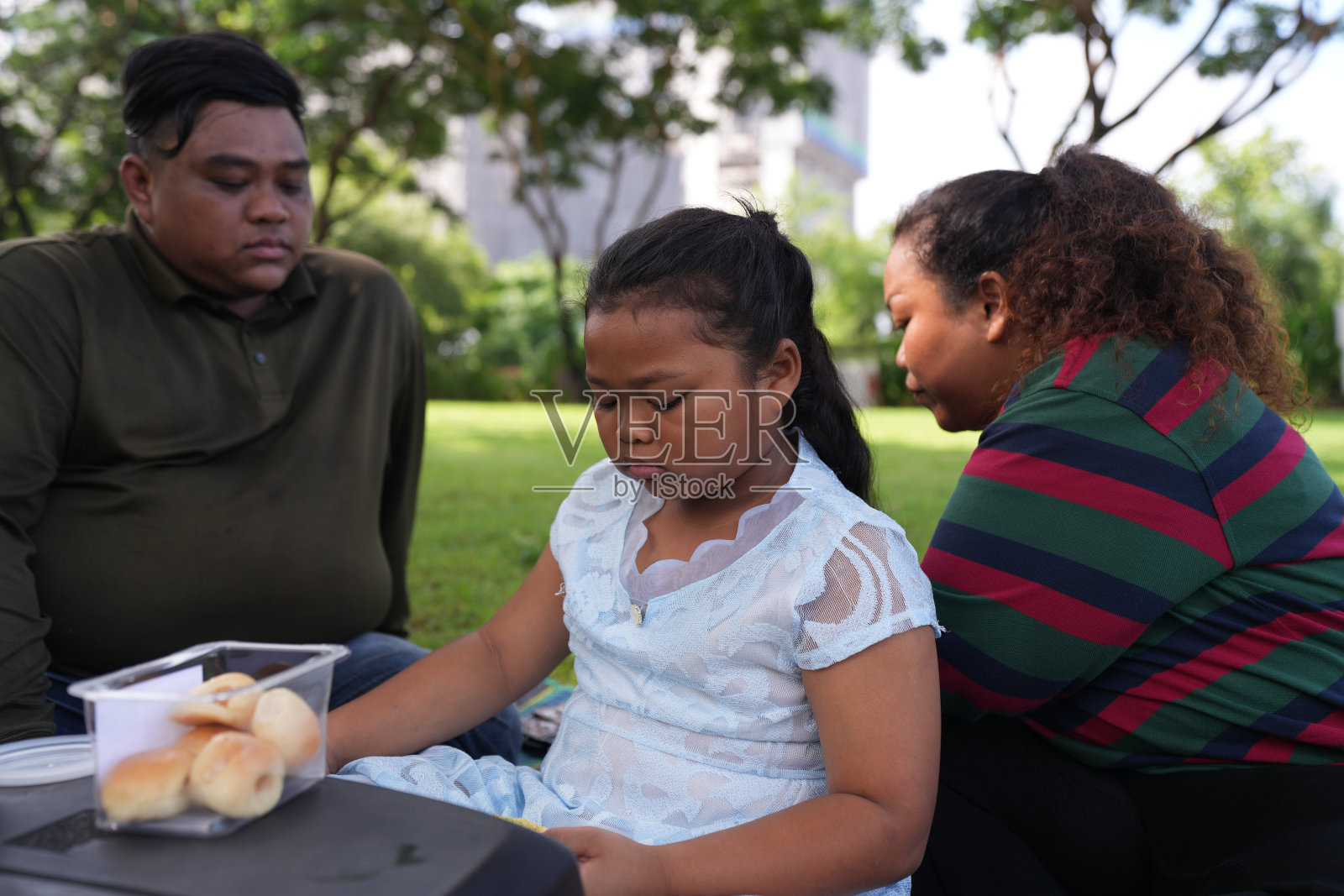 一个又大又胖的亚洲家庭,爸爸、妈妈、女儿在公园里野餐,他们坐着吃零食。他们坐着做作业,互相送花。他们很开心。照片摄影图片