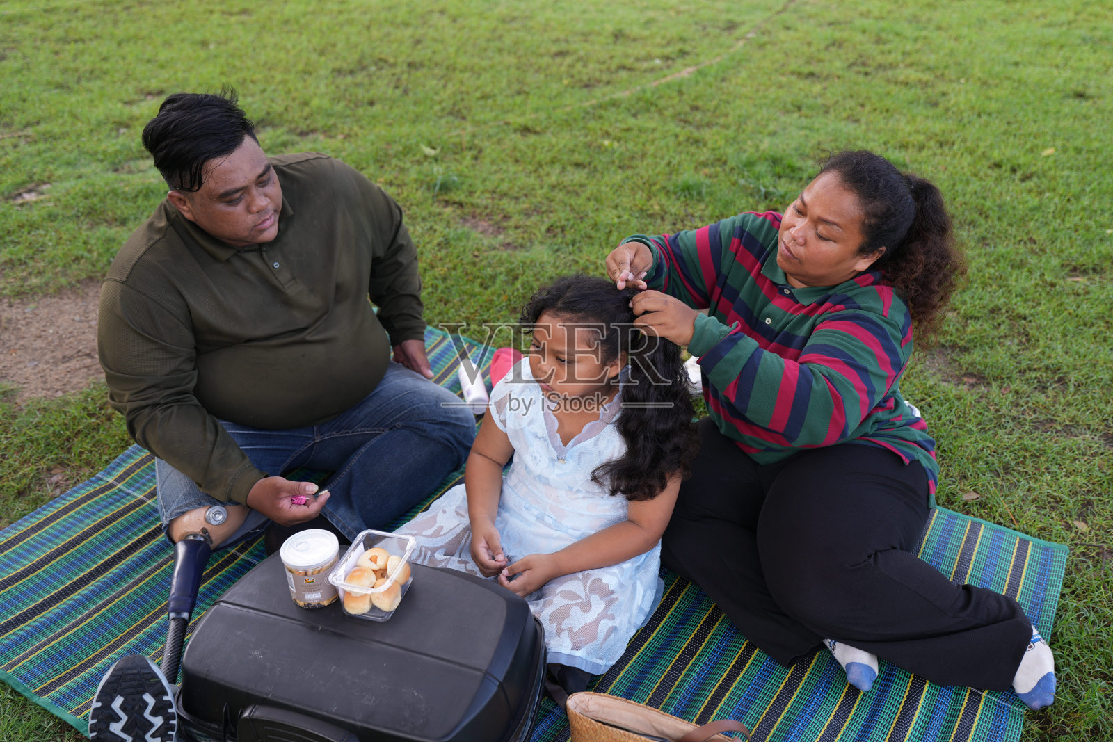 一个又大又胖的亚洲家庭,爸爸、妈妈、女儿在公园里野餐,他们坐着吃零食。他们坐着做作业,互相送花。他们很开心。照片摄影图片