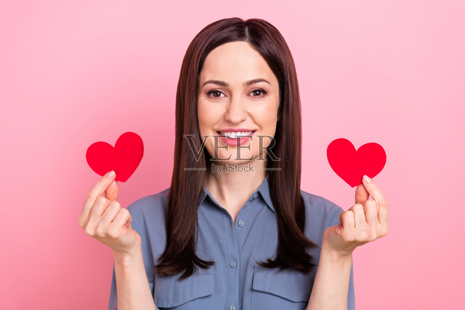 开朗积极的年轻女经理的肖像,穿着蓝色时髦的衬衫,拿着两张纸爱心明信片,孤立在粉红色的背景上照片摄影图片
