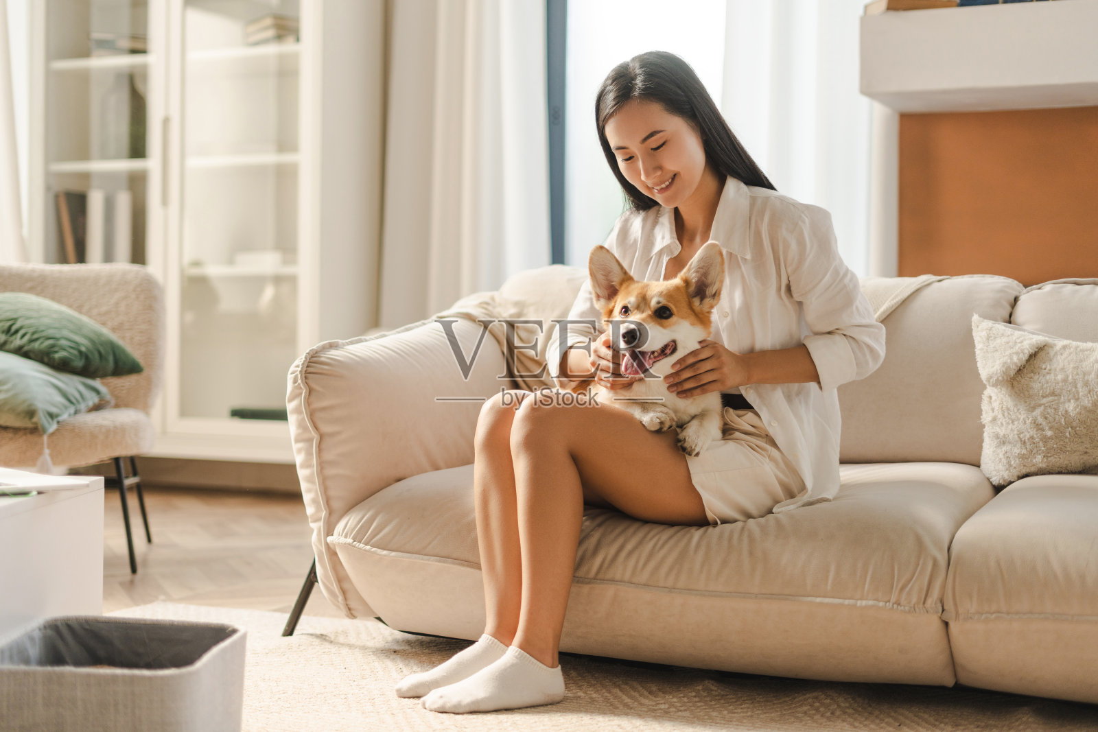 年轻,微笑的亚洲女人放松地躺在沙发上,抱着她的宠物威尔士柯基彭布罗克狗照片摄影图片