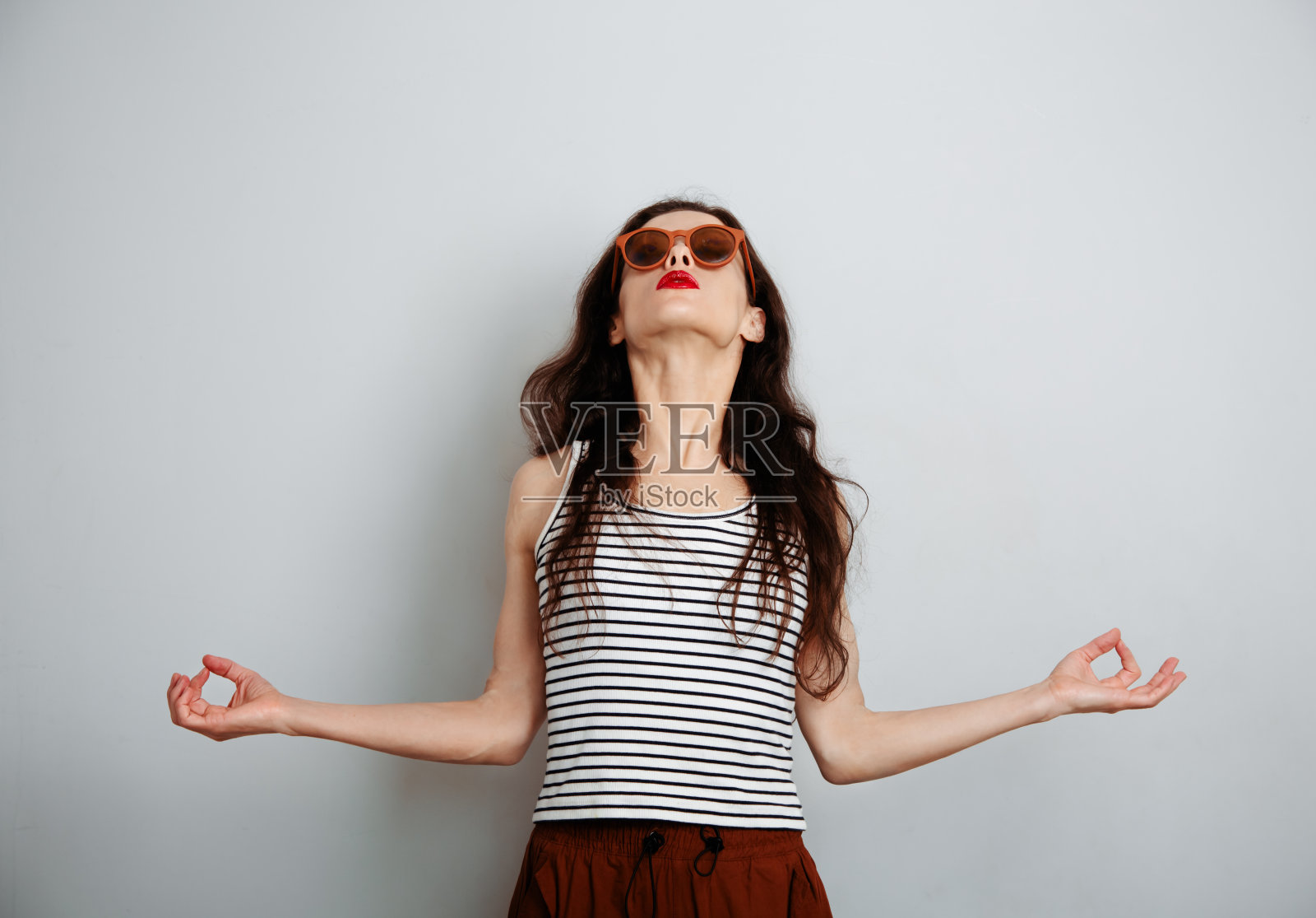 美丽的化妆师做瑜伽冥想禅宗的手指在休闲上衣,红色的口红和长发,在蓝色工作室背景的时尚太阳镜。特写镜头照片摄影图片