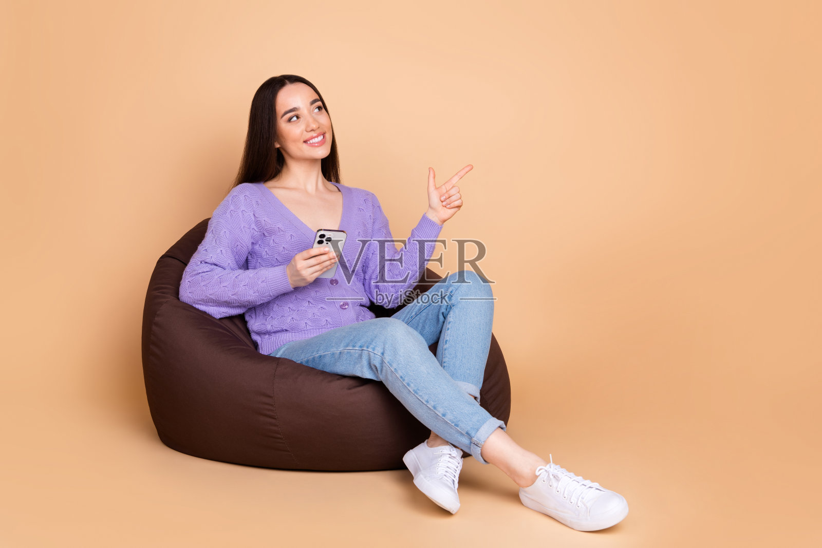 漂亮年轻女子的全身肖像,智能手机显示手指空白,穿着开衫孤立在米色背景上照片摄影图片