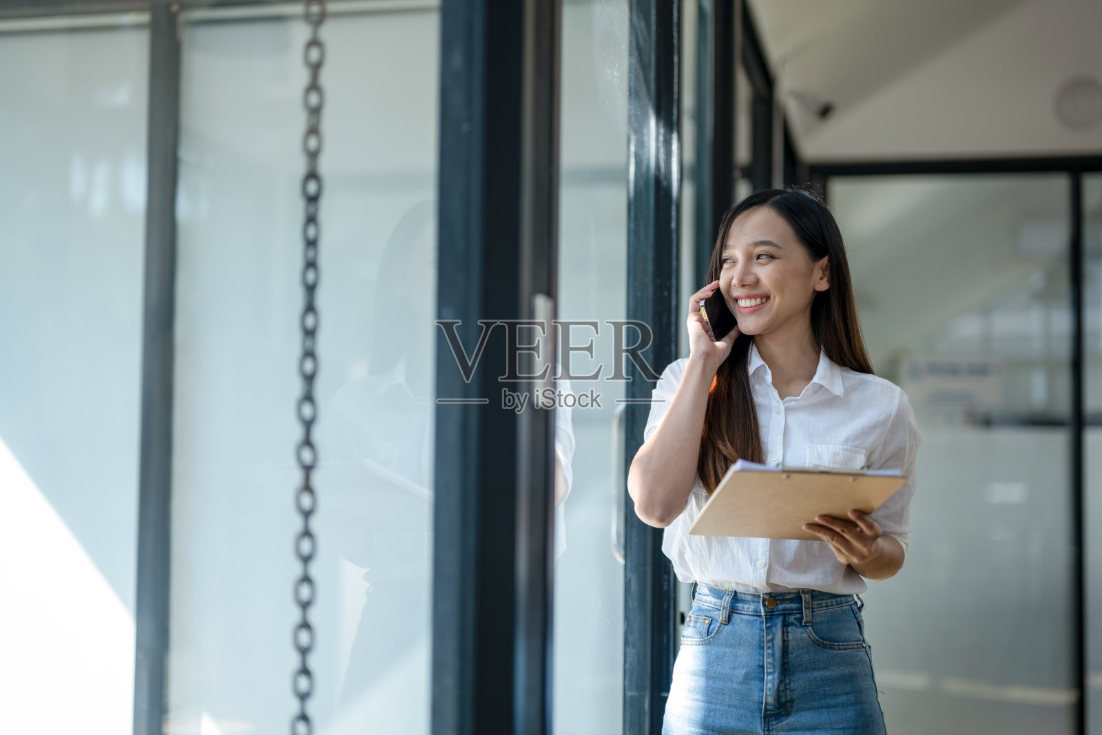 在光线充足的现代办公环境中,微笑的年轻亚洲女商人拿着写字板打电话。照片摄影图片