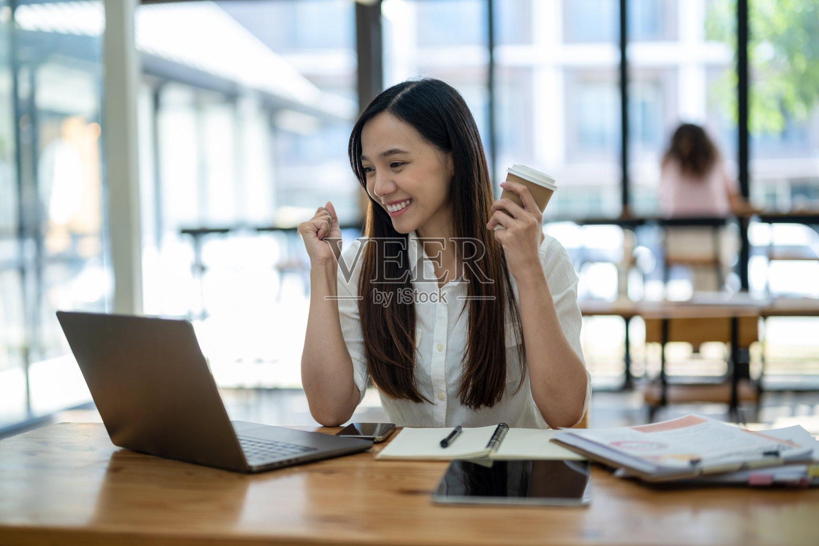 在明亮的办公环境中,一位友好的亚洲女商人拿着咖啡杯,在笔记本电脑上进行视频通话时向她打招呼。照片摄影图片