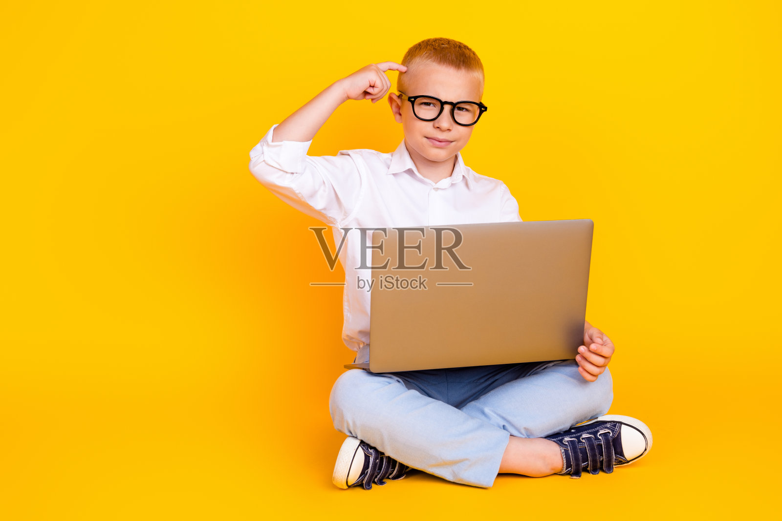 聪明聪明的男孩穿着白衬衫坐在地板上摸着头的照片决定孤立在黄色的背景上照片摄影图片