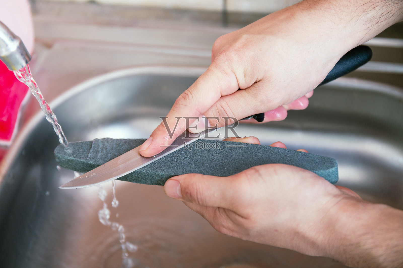 特写:一个男人的手在水流下小心地在磨刀石上磨刀。家居家居磨刀。照片摄影图片