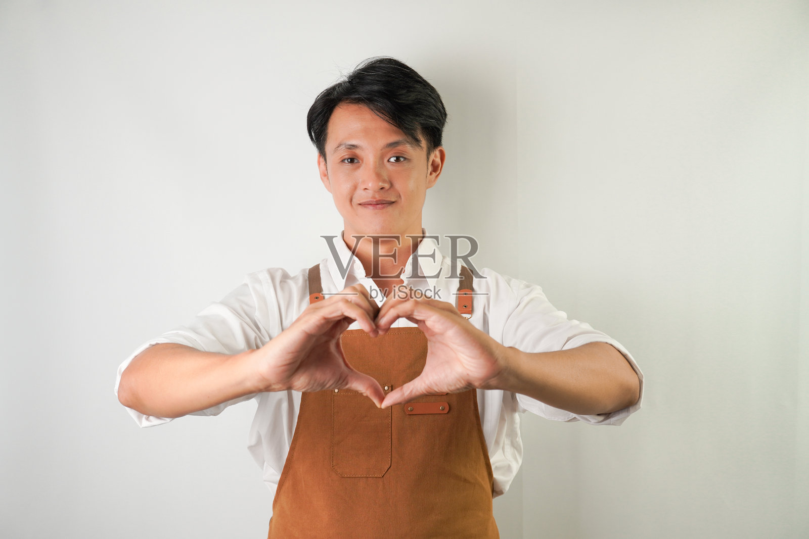 身穿卷起白色袖子的衬衫和棕色围裙,用手指做出爱心手势的友善亚洲青年男子。隔绝在白色背景上。照片摄影图片