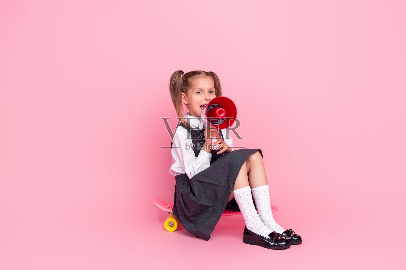 小女生的全身照片骑滑板宣布扩音器穿着时尚的制服孤立在粉红色的背景照片摄影图片