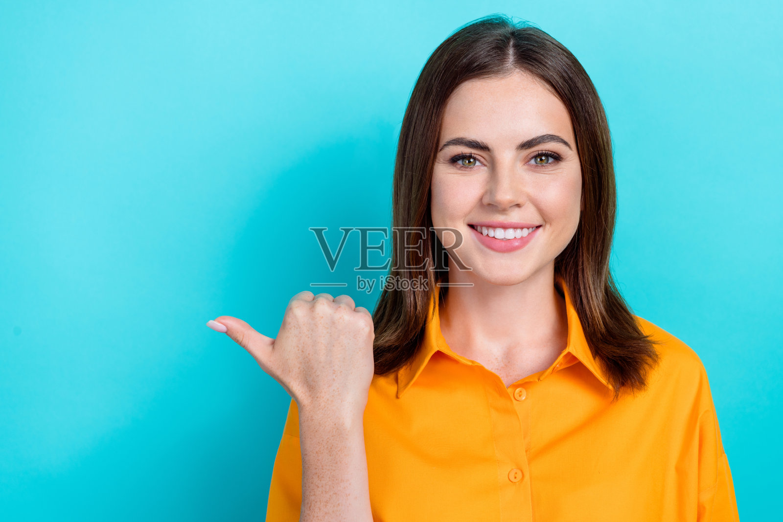 照片中漂亮可爱的女人穿着橙色的衬衫,手指着空白的空间,孤立的蓝绿色背景照片摄影图片