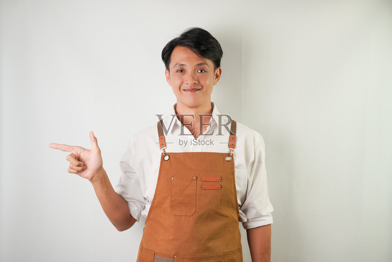 穿着卷起的白袖衬衫,系着棕色围裙的兴奋的年轻亚洲人正带着微笑的表情指着旁边。隔离在白色背景上照片摄影图片