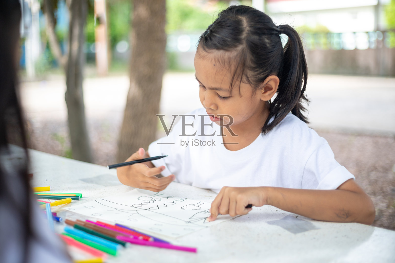 年轻的女孩们坐在桌子旁,桌上放着马克笔和涂色书。他们在一起画画和涂色,享受他们在公园的时光。乐趣和创造力的概念照片摄影图片