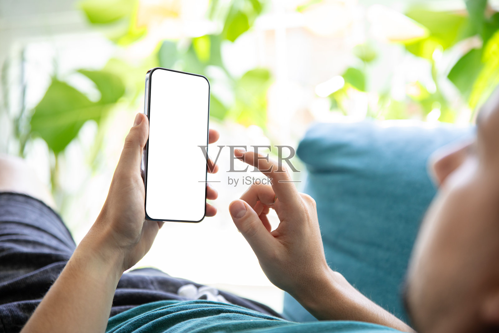 一个人拿着空白屏幕的智能手机,在明亮的绿色环境中躺在沙发上放松。完美的图像呈现移动应用程序,用户界面,或设计模板。照片摄影图片