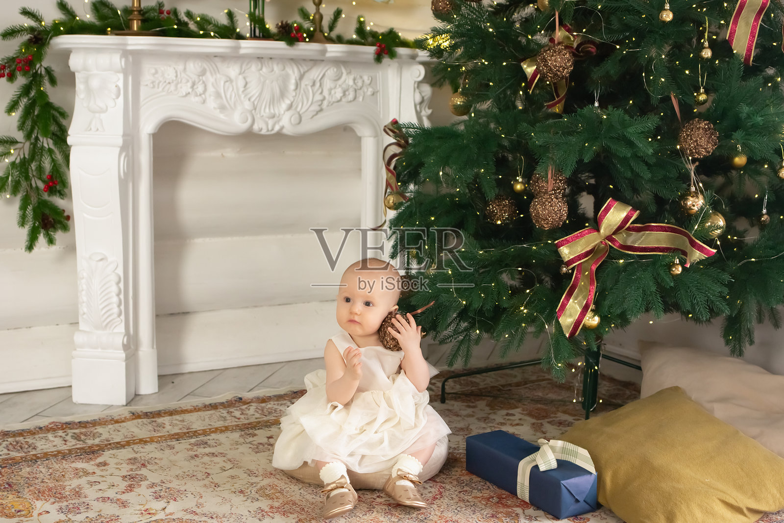 在为新年装饰的客厅里,一个穿着漂亮的白色连衣裙的小女孩坐在圣诞树旁边照片摄影图片