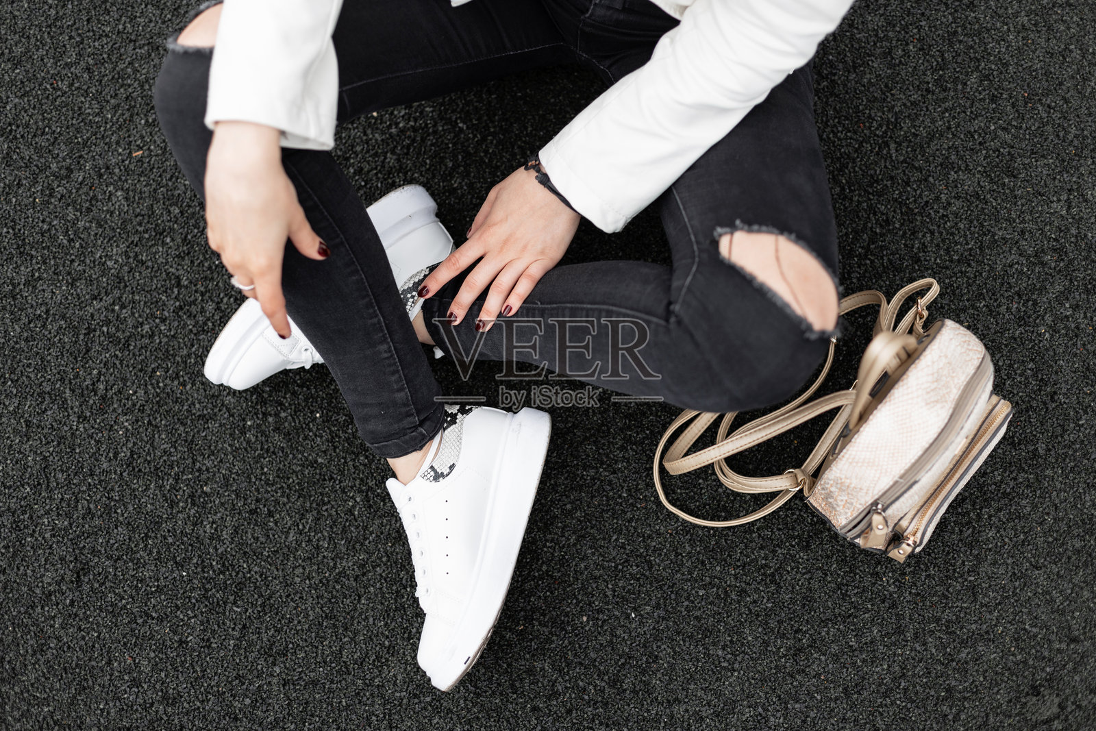 一名年轻女子穿着白色夹克和时尚的黑色破洞牛仔裤,背着金色时髦背包,穿着时尚的皮运动鞋,坐在柏油路上。日常外观的细节。从上面看。照片摄影图片