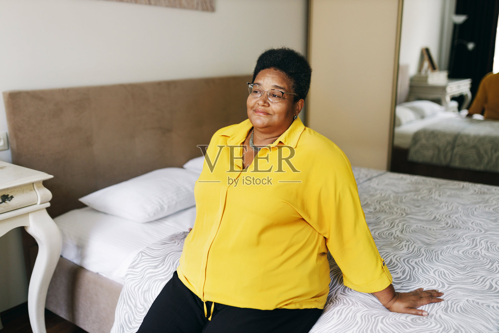 穿着黄色衣服,戴着眼镜的大码非裔美国祖母坐在床上,双手倚着,脸上带着沉思和积极的表情,回忆着她生命中最美好的回忆照片摄影图片