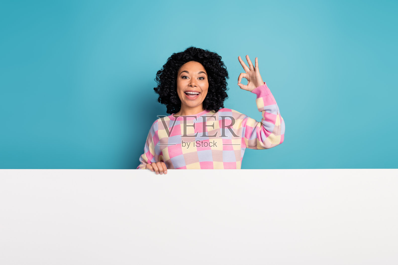 迷人的年轻女子站在白色的空地后面,穿着时髦的粉红色格纹衣服,与蓝色的背景分开照片摄影图片