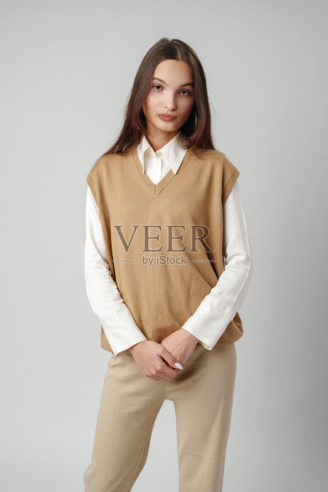 一头棕色长发,身穿白衬衫和棕色毛衣的年轻女子在一堵灰色的墙前看起来很悲伤照片摄影图片