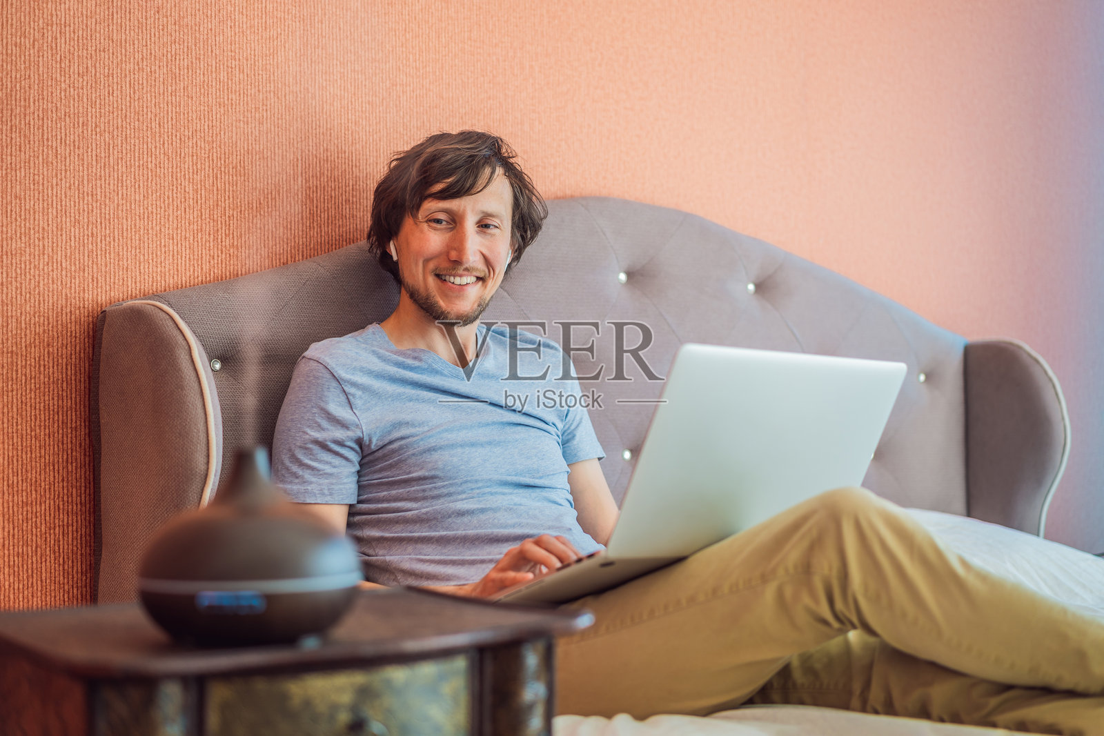 男人坐在床上,用笔记本电脑工作。床边的芳香扩散器照片摄影图片