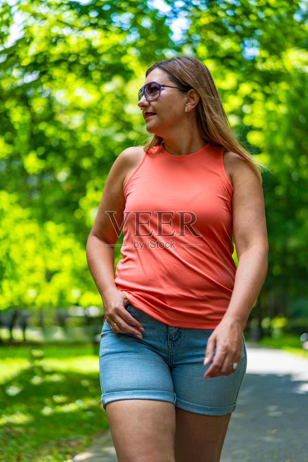 年轻的大码金发女子的肖像,穿着杏黄色的运动衫和牛仔短裤,戴着太阳镜,在阳光明媚的日子里,在公园的绿色背景下散步,看着旁边。照片摄影图片