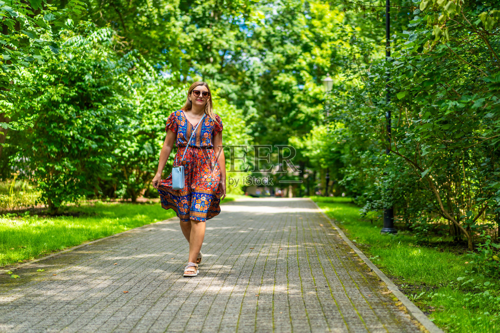 美丽的金发女子穿着五颜六色的碎花飘逸的连衣裙,在阳光明媚的日子里,在城市公园的绿色背景下散步。前视图照片摄影图片
