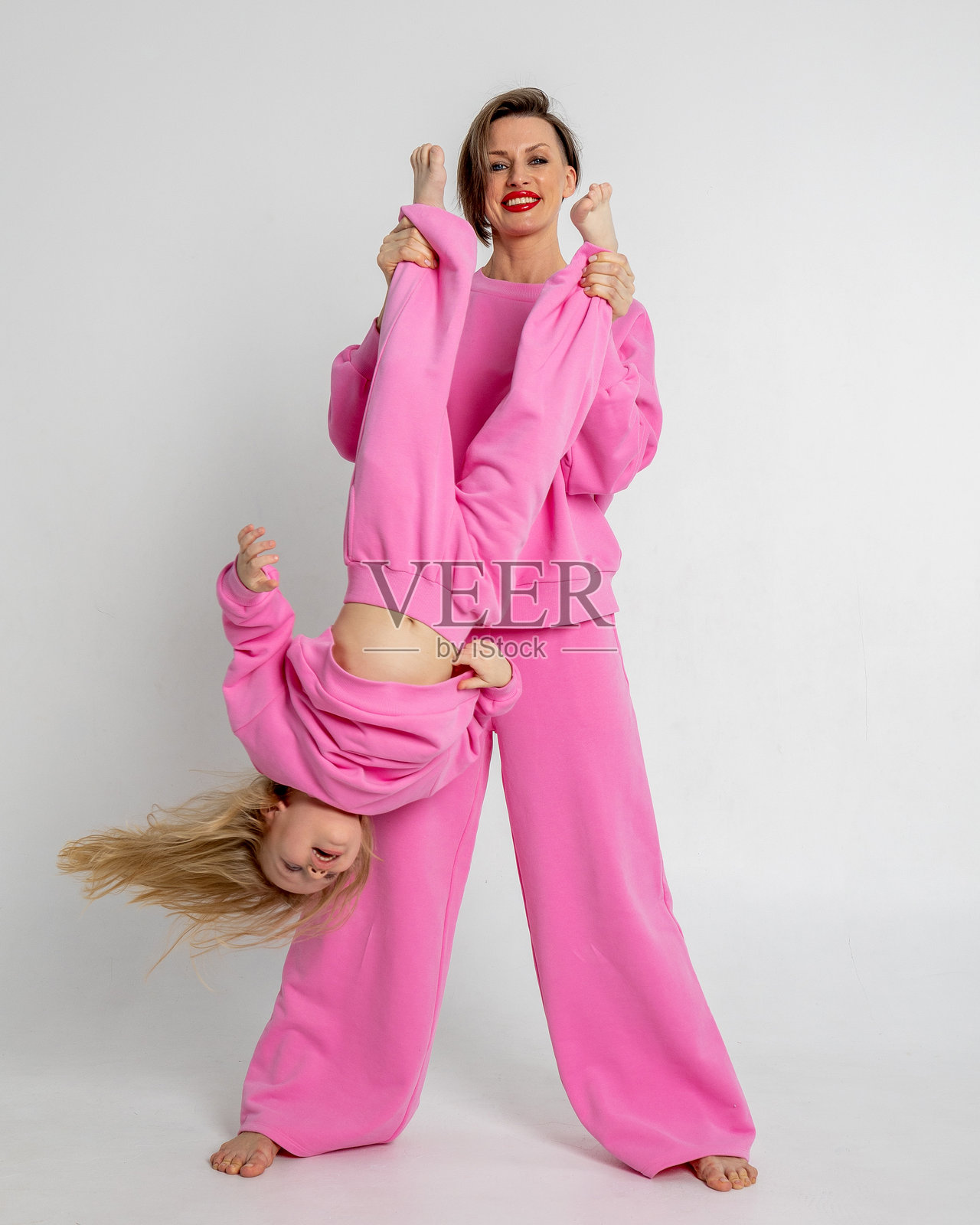 年轻的母亲和小女儿手牵着手走着。穿着白色背景的粉色西装,工作室。广告理念积极和服装照片摄影图片