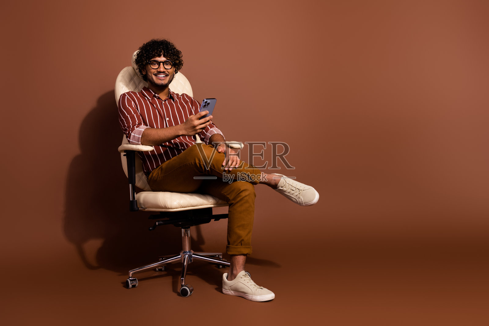 漂亮年轻人的全身肖像,坐在椅子上,拿着智能手机,穿着条纹衬衫,孤立在棕色背景上照片摄影图片