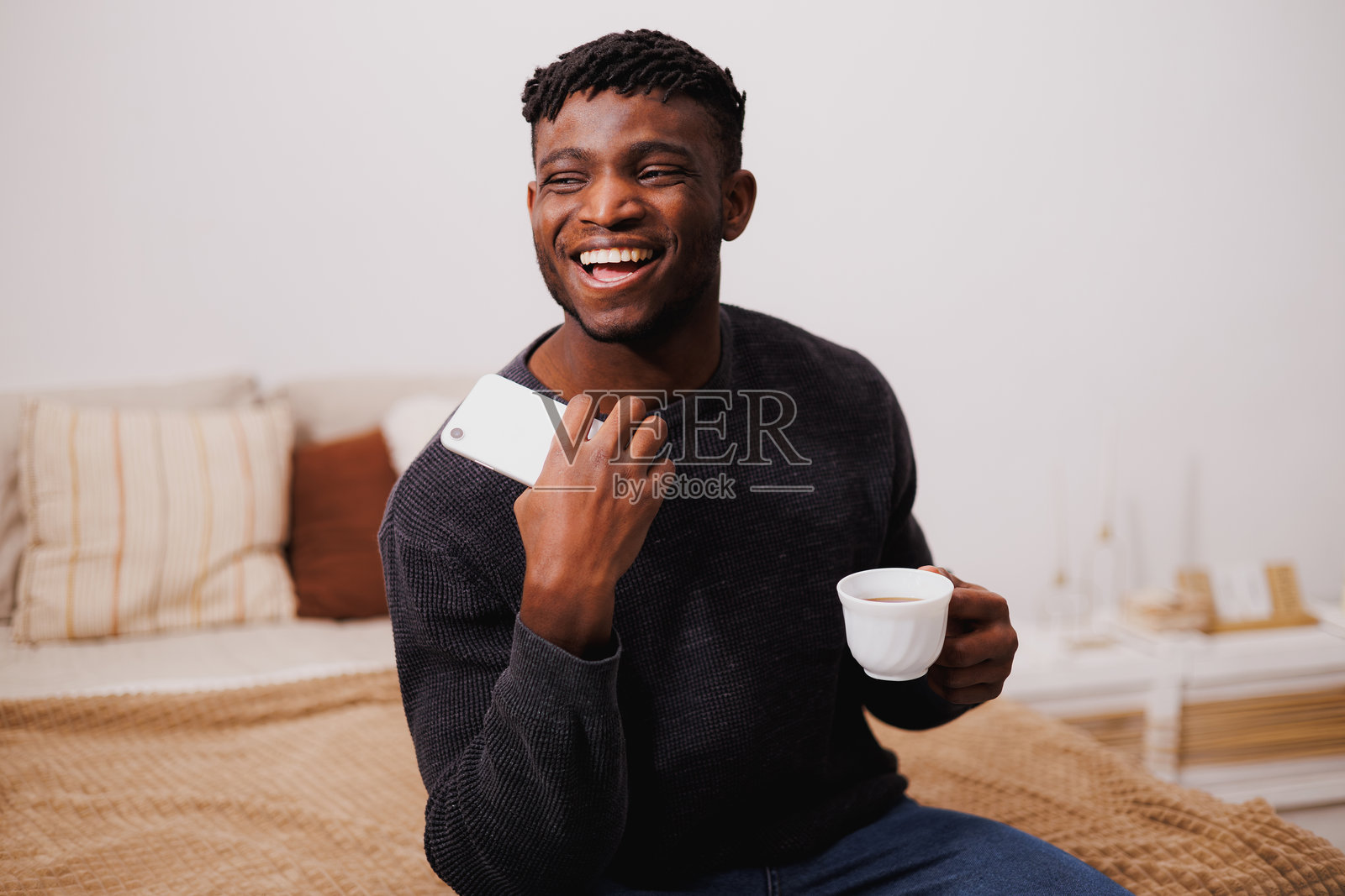 快乐的非裔美国人穿着套头衫,拿着智能手机和咖啡放在床上照片摄影图片