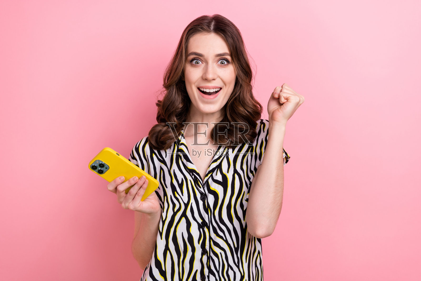 欣喜若狂的年轻女孩的照片举起拳头庆祝玩得开心玩电话在线赌博彩票棕色波浪的头发孤立在粉红色的背景照片摄影图片