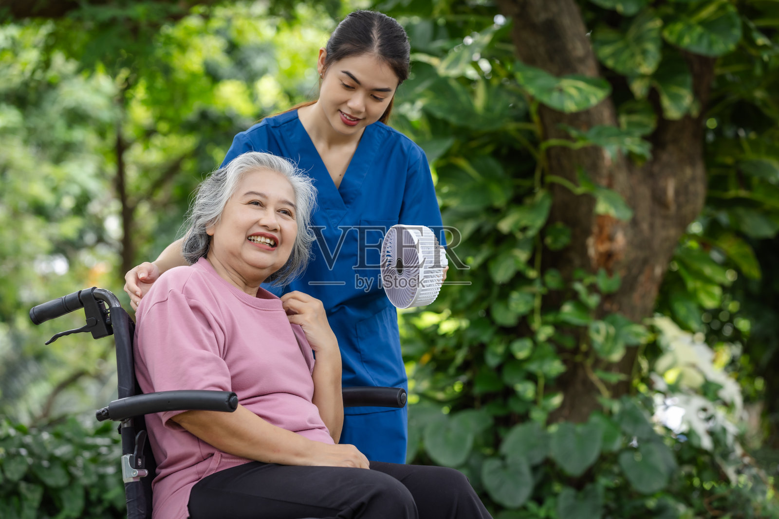 一位坐在轮椅上微笑的老妇人在花园里享受着由一位关怀的护士拿着的便携式风扇吹来的凉风照片摄影图片