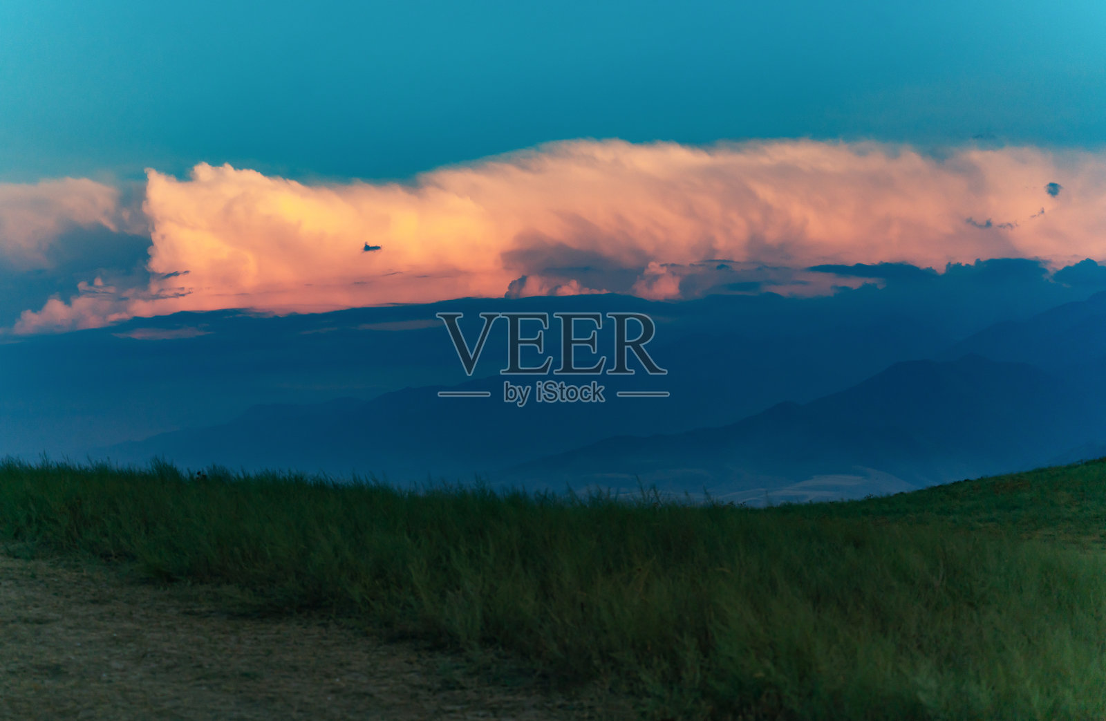 云是一个笑脸。从长满绿草的小山上看到一片明亮的大云,被夕阳的光线染成颜色,远处是高山。云朵的右边露出了一丝微笑。水平的照片。照片摄影图片