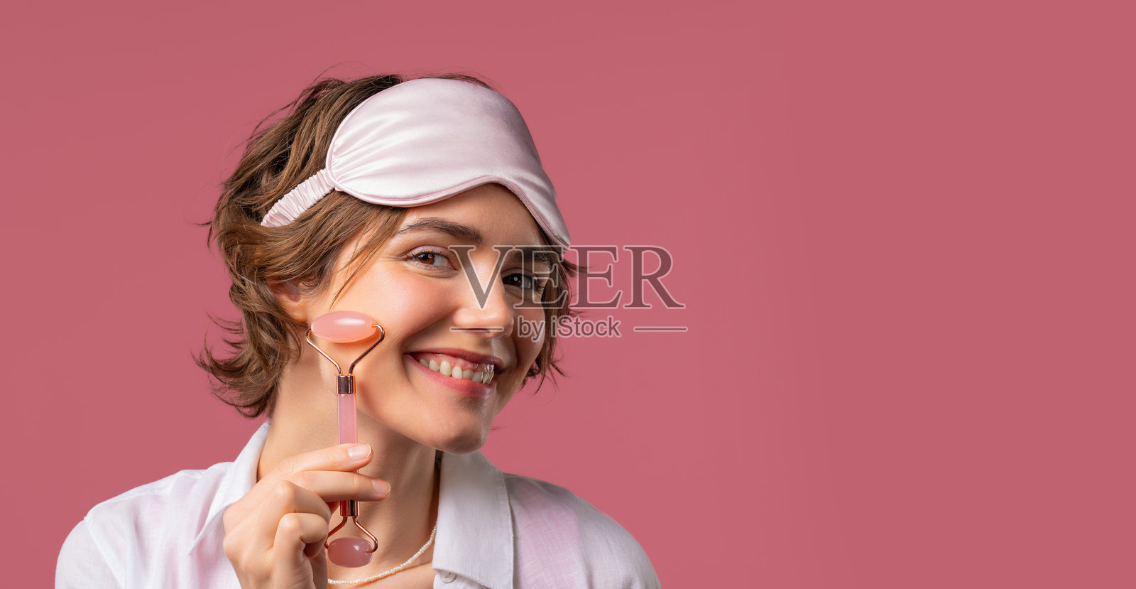 年轻女子与玫瑰石英石辊在粉红色的背景。面部自我护理,早晨美容仪式,美容,抗衰老和抗皱治疗照片摄影图片