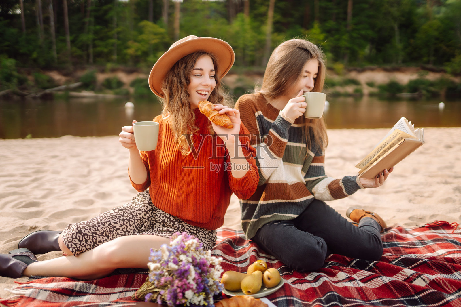 在一个阳光明媚的下午,两个朋友在河边野餐,他们拿着鲜花,在舒适的毯子上分享零食。照片摄影图片