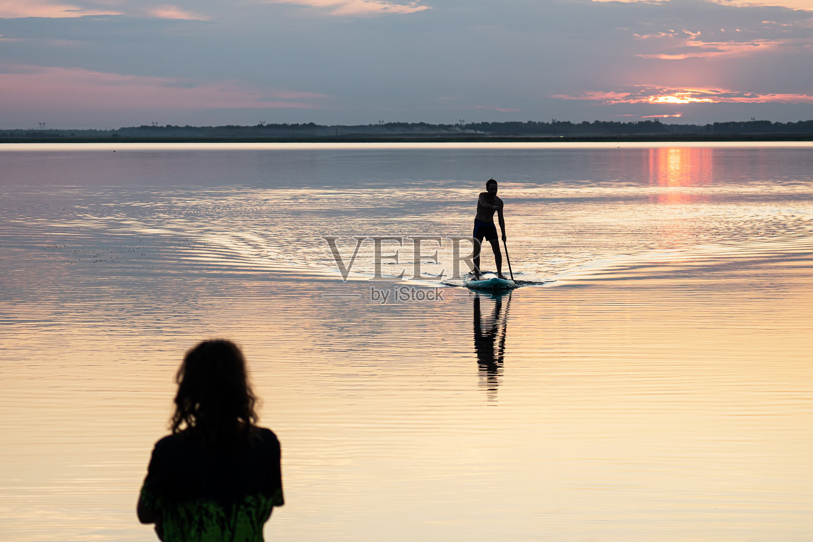 夕阳中独自一人的男性划桨者的侧影。在海湾里划桨。日落时分,一个年轻人在湖面上用立式桨板划桨。照片摄影图片