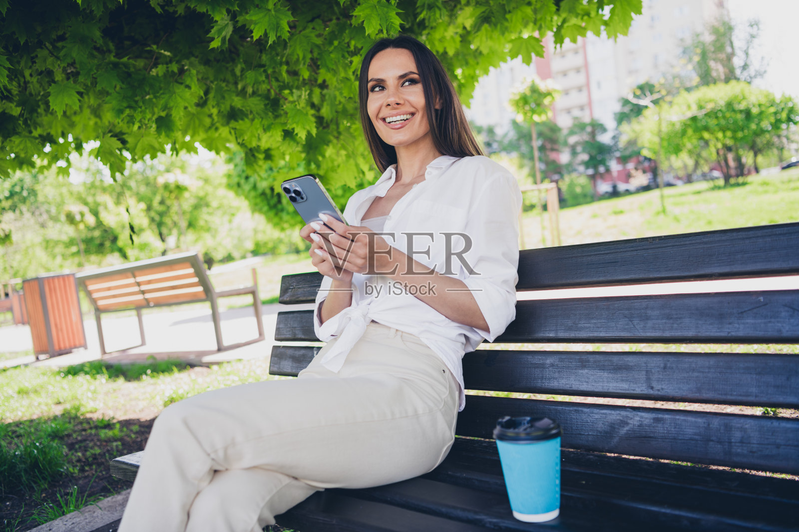 迷人的年轻开朗的女孩的照片,坐在长凳上,咖啡,拿着装置,穿着白色的衣服在户外玩耍照片摄影图片