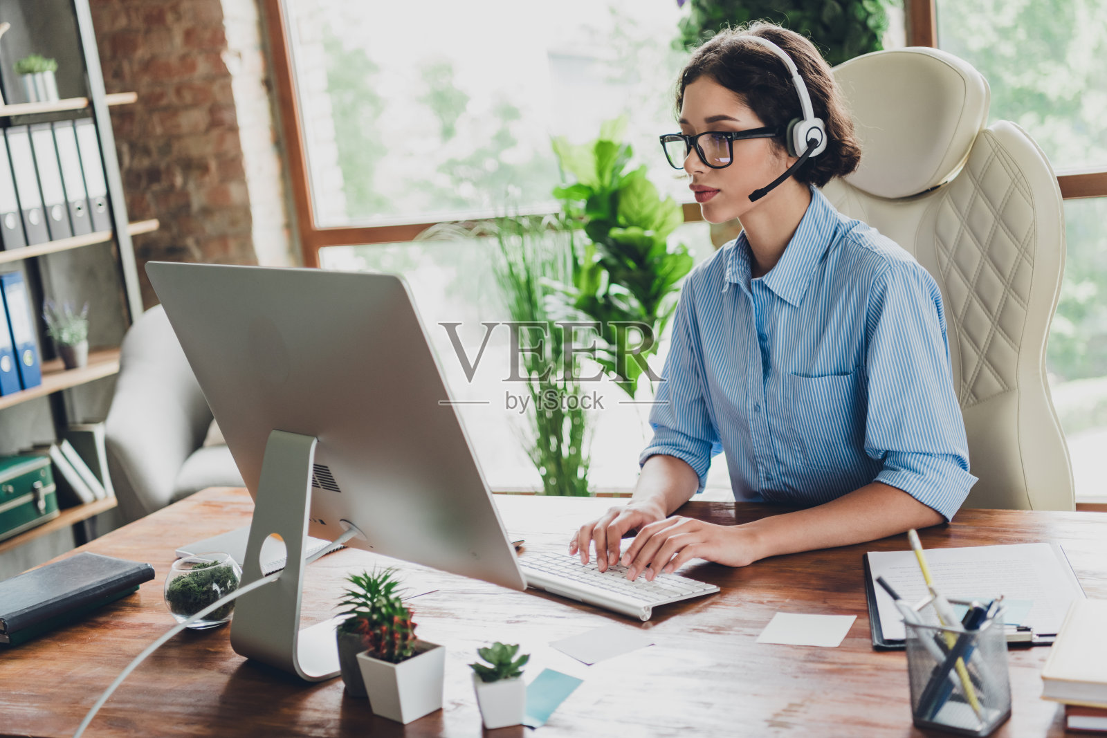 甜美迷人的女士助理的照片穿衬衫,眼镜,耳机短信现代设备在室内工作场所工作站照片摄影图片