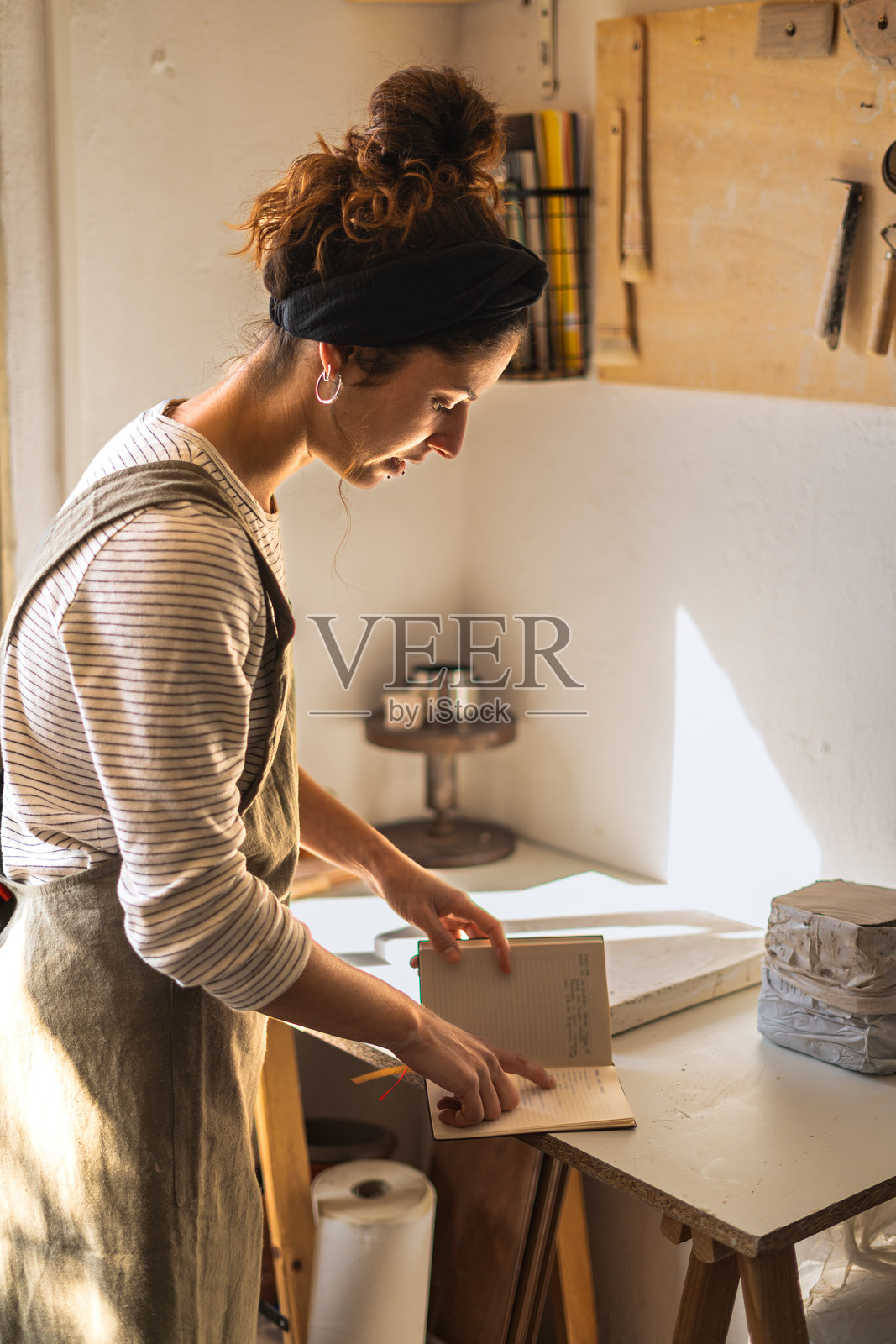在一间温暖的陶艺工作室里,一位系着围裙的艺术家仔细地检查着她的笔记,工作室周围摆满了她的专业工具照片摄影图片