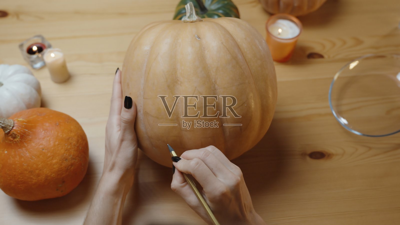 木桌上放着蜡烛的南瓜,女人在南瓜上画了一张脸的草图,刻了一个南瓜灯。前视图。观点。照片摄影图片