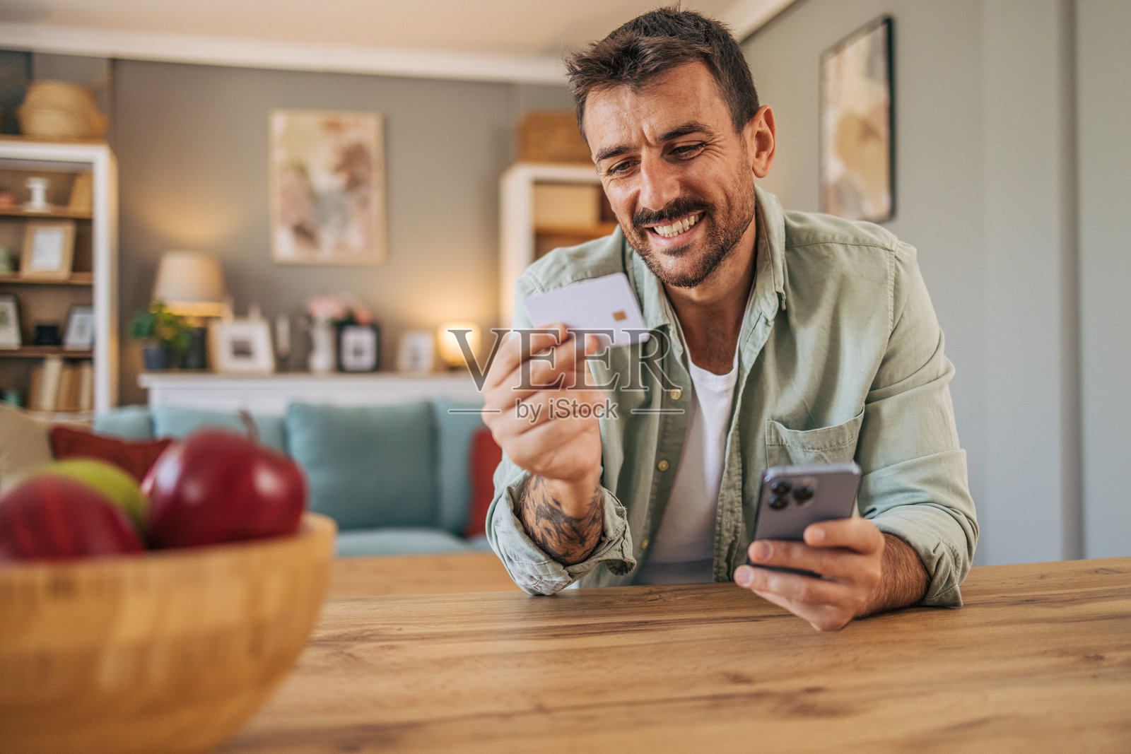 一位英俊、开朗的中年男子坐在家里客厅的桌子旁,正在使用智能手机。他用信用卡在网上支付账单或网上购物。照片摄影图片