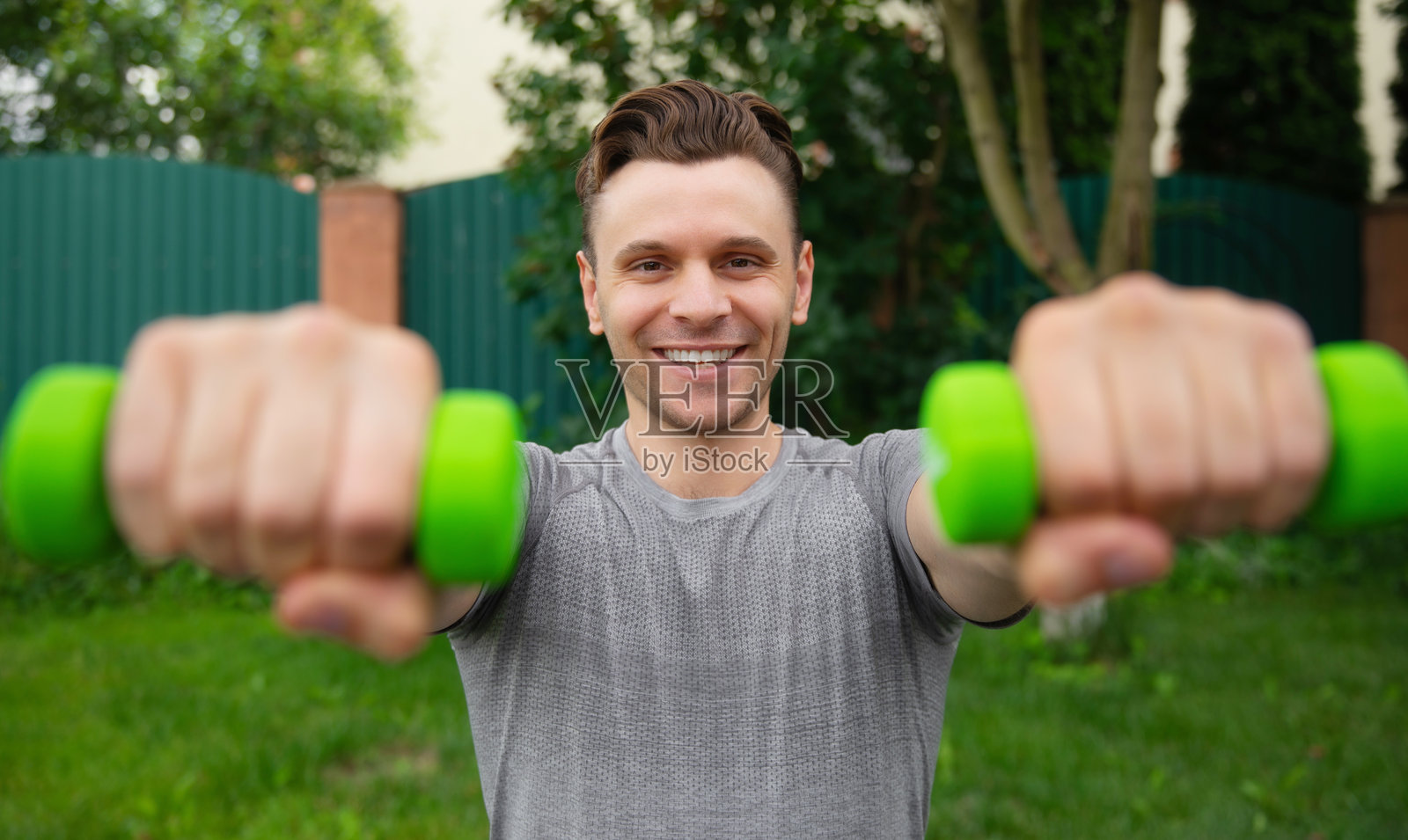 微笑的男子在户外锻炼时双手举着两个哑铃。画面捕捉了一个快乐和对健身的投入时刻,强调了在生机勃勃的自然环境中进行力量训练。照片摄影图片