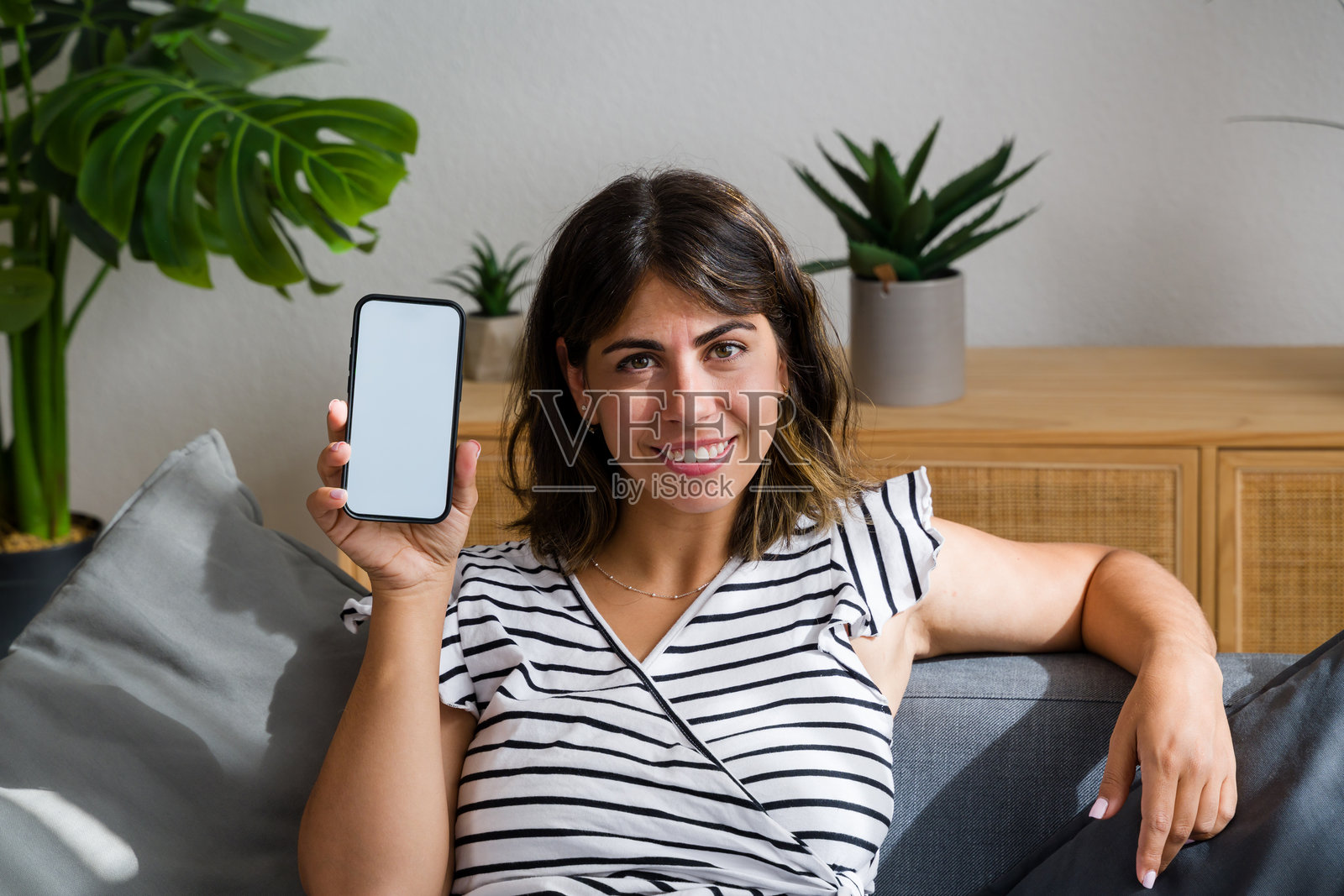 女人微笑着坐在家中办公室的沙发上,展示着一部空白屏幕的智能手机。照片摄影图片