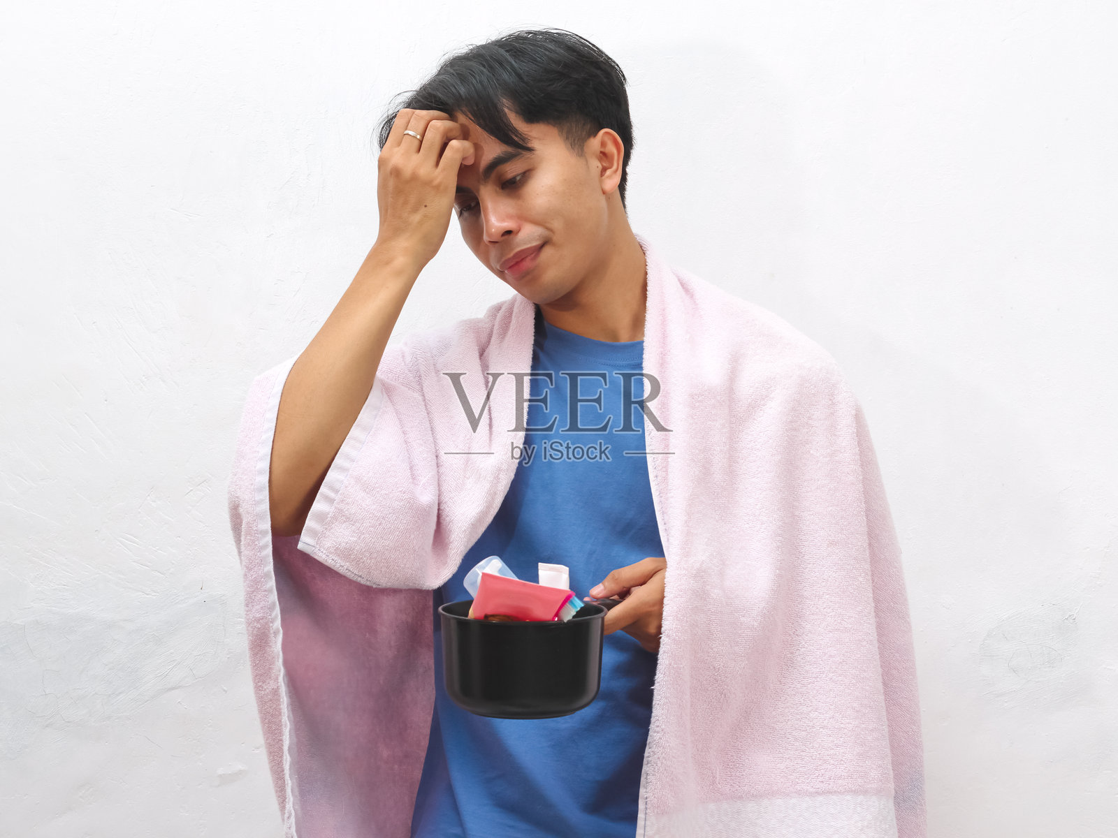 一个疲惫的亚洲男人拿着毛巾和洗漱用品,看起来很懒,刚刚醒来,背景是白色的。照片摄影图片