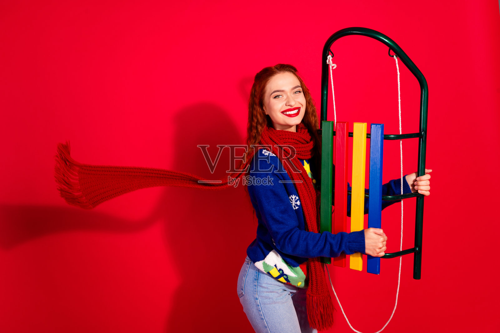 可爱的年轻女士手持雪橇,围着长围巾,穿着时尚的蓝色圣诞服装,在红色背景下庆祝冬季假期。照片摄影图片