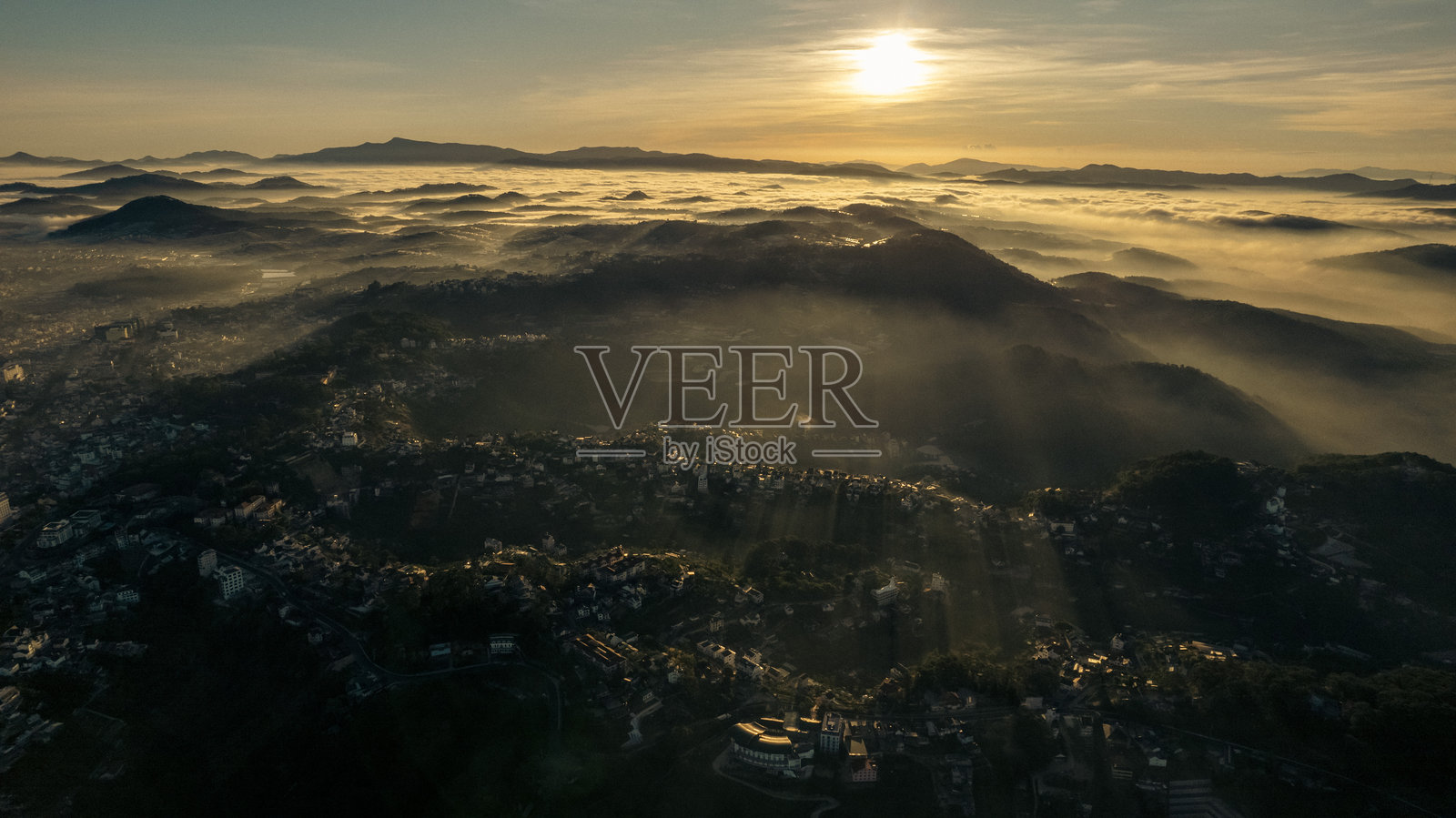 日出时分的大叻市鸟瞰图,展现了城市景观与自然美景的结合,建筑坐落在郁郁葱葱的山丘和雾气弥漫的山谷之间,沐浴在金色时光的光辉下。照片摄影图片