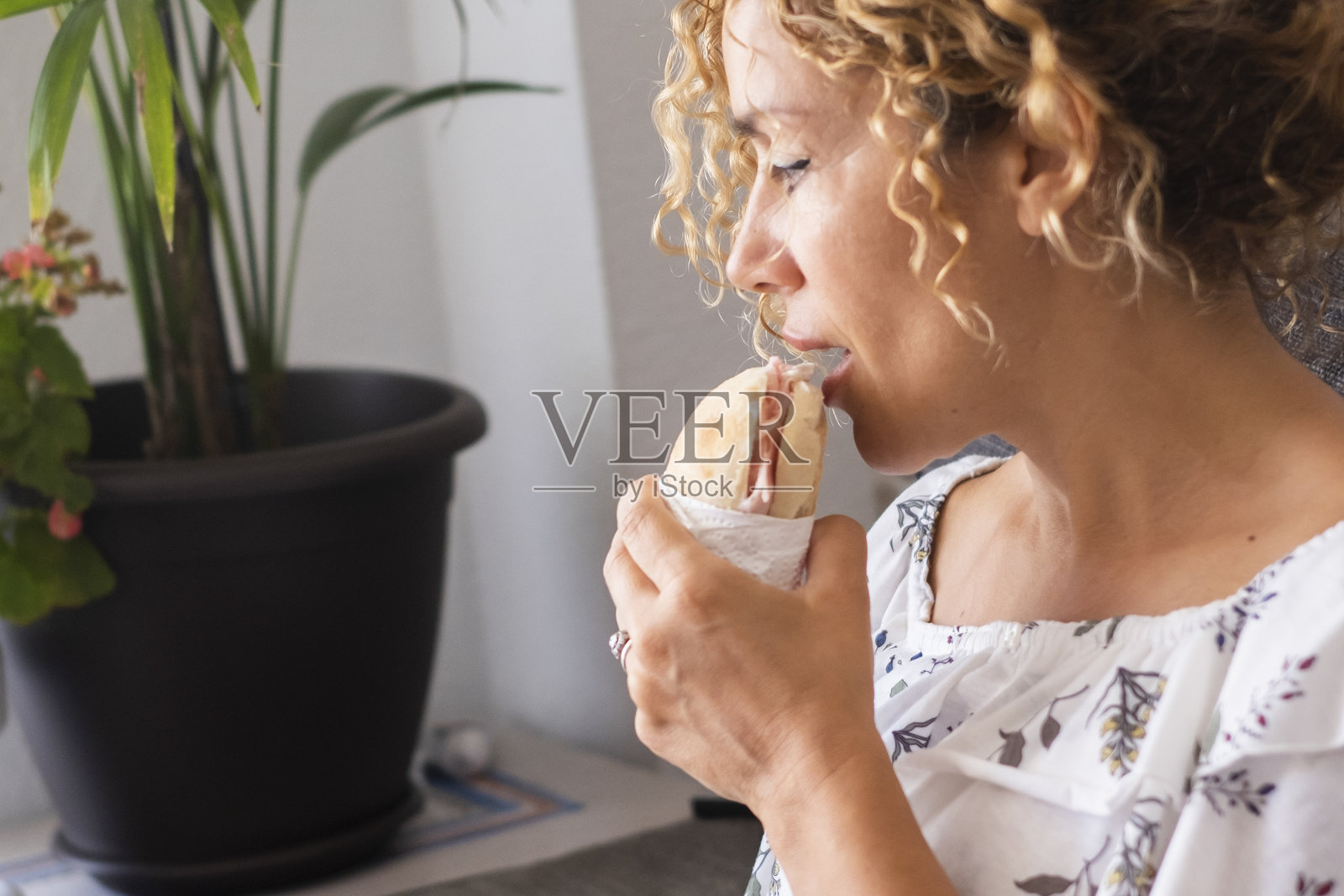 一位成年女性在家中享用三明治的特写镜头,享受着快速且有些不健康的午餐。轻松的室内环境突显了简单美味餐点的便利与放纵。照片摄影图片