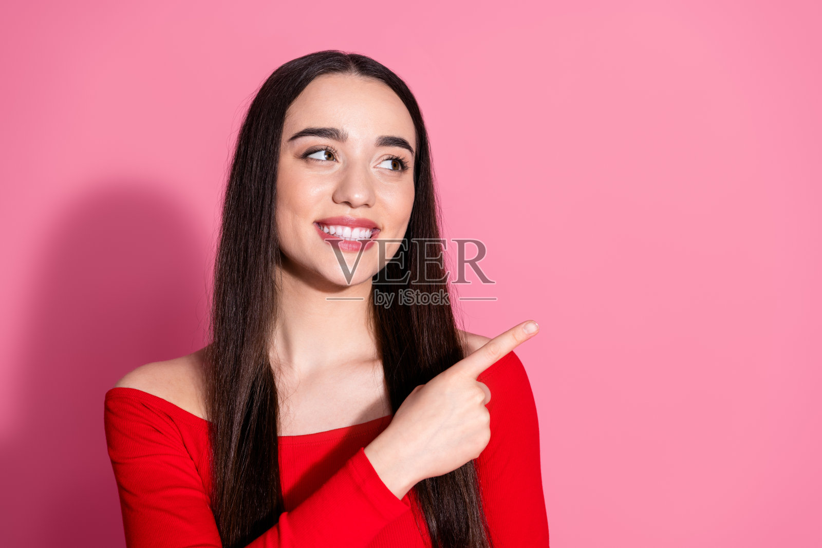 穿红色上衣的可爱年轻女孩用手指指向粉色背景上的空白区域照片摄影图片