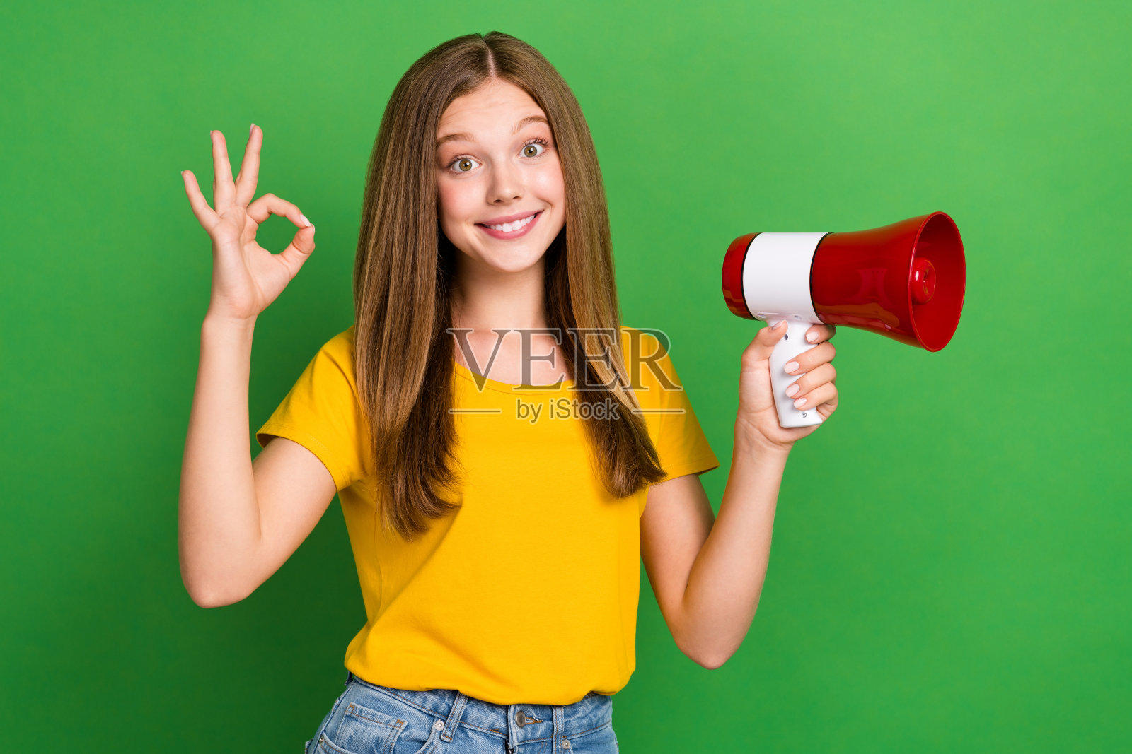 可爱的快乐女孩手持扬声器,手指做出“好”的手势,背景为绿色。照片摄影图片