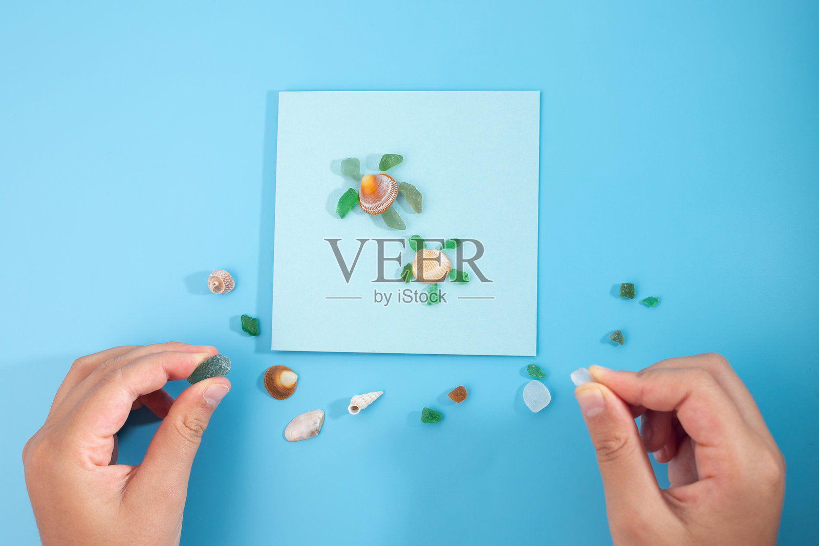 蓝色背景上的平铺图,展示浅蓝色纸张、海贝壳、绿色玻璃碎片,手放置贝壳,营造海滩场景,手工艺品,儿童艺术项目,自然材料,DIY。照片摄影图片