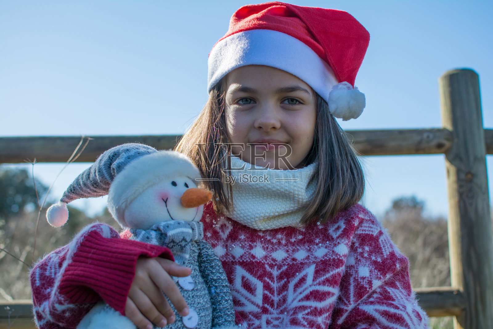 戴着圣诞帽和红色圣诞毛衣的快乐女孩在户外抱着一个雪人玩具照片摄影图片