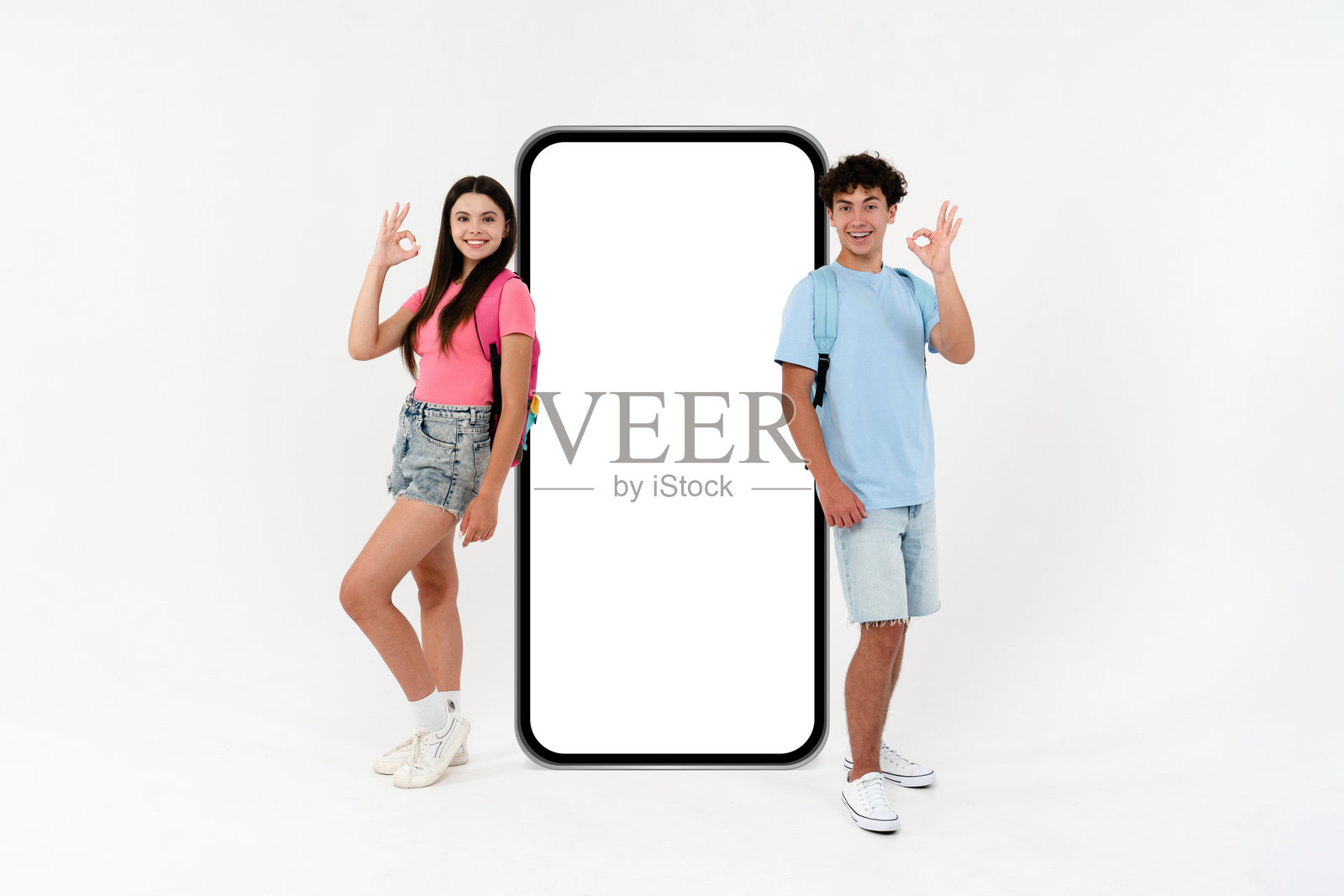 高中生男孩女孩全身照,穿着休闲,做出“好的”手势,站在一个大模型手机旁,手机屏幕区域为空,背景为白色。照片摄影图片